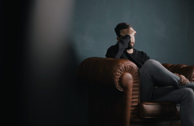 A male employee who is experiencing workplace bullying sits alone on a sofa with his hands over his eyes.